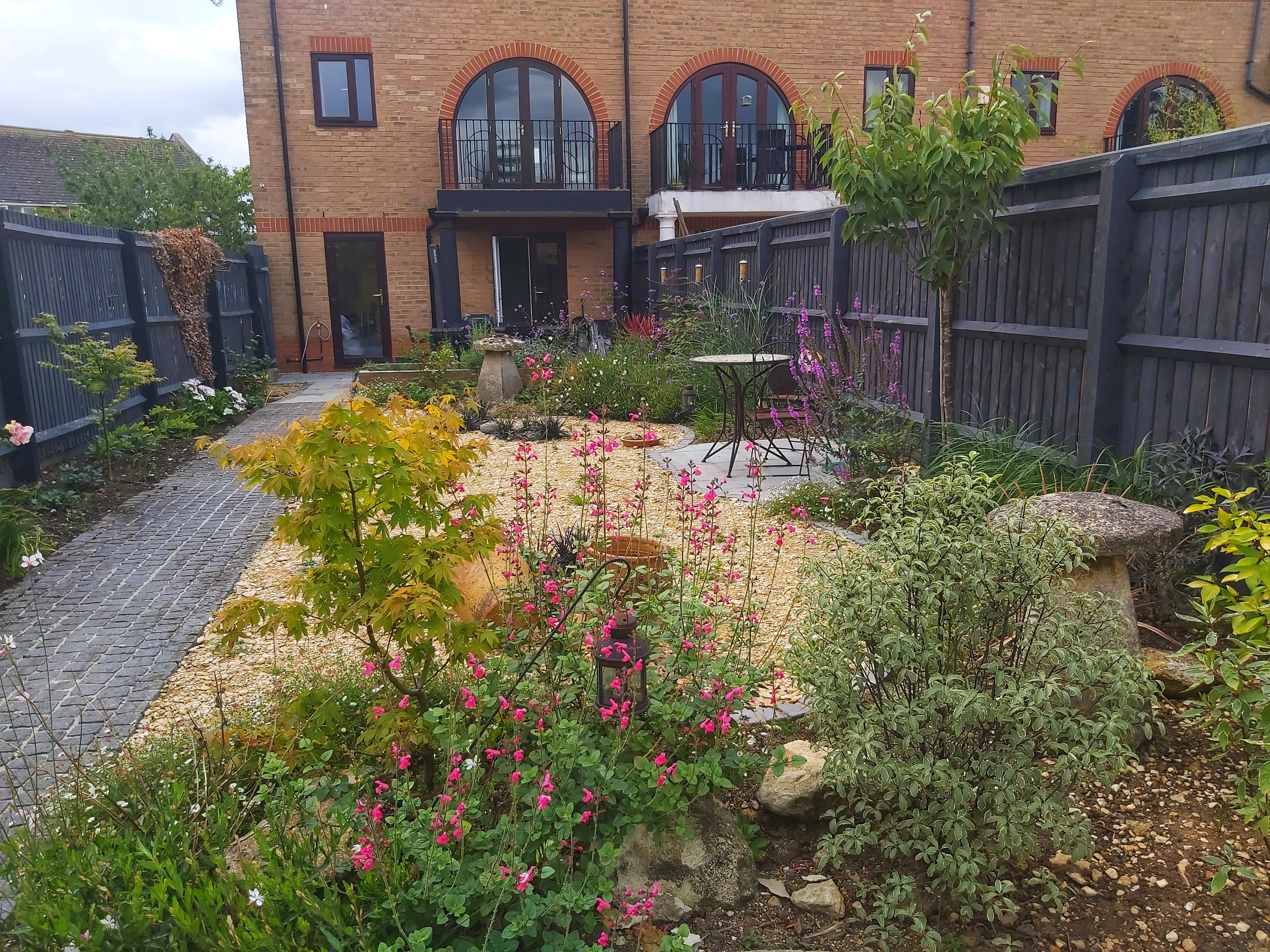 The Low-Maintenance, Bee-Friendly Garden — After The same view of the house. The grass has been replaced with a bistro set on gravel, surrounded by bright flowerbeds