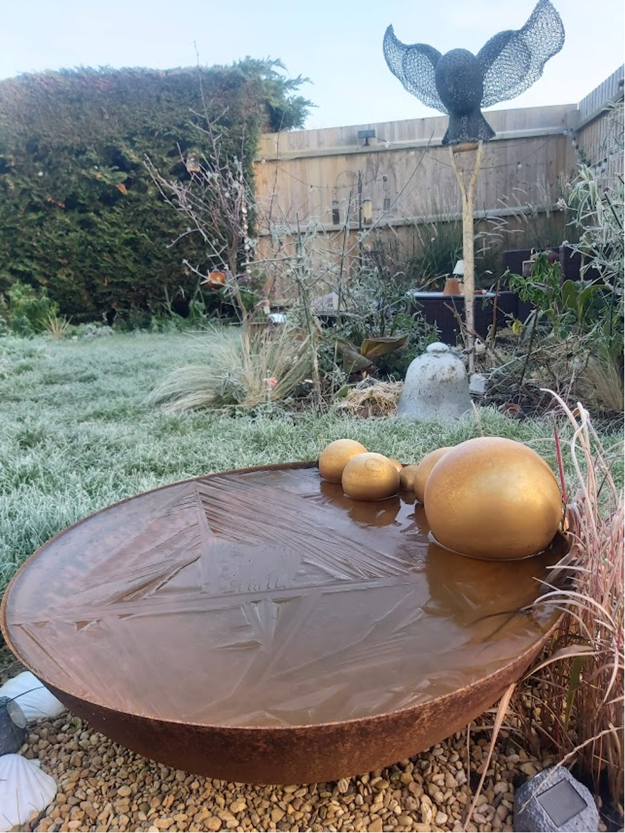 A frozen-over pond in a metal bowl on a frosty lawn. A sculpture of an owl made from wire looks over